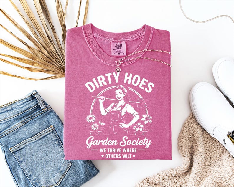 May include: A pink t-shirt with the words "Dirty Hoes Garden Society" and a graphic of a woman holding a gardening tool. The shirt is paired with blue jeans, white sneakers, and a necklace.