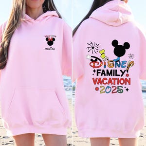 May include: Pink Disney Family Vacation 2025 hoodie with a Mickey Mouse graphic and fireworks. The front of the hoodie has a smaller Mickey Mouse graphic with the text "Disney 2025" and "Monica".