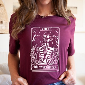 May include: A maroon t-shirt with a white graphic of a skeleton in a tarot card design. The card reads "The Overthinker".