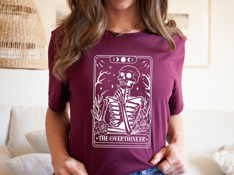 May include: A maroon t-shirt featuring a white graphic of a skeleton in a tarot card design. The card reads "The Overthinker".