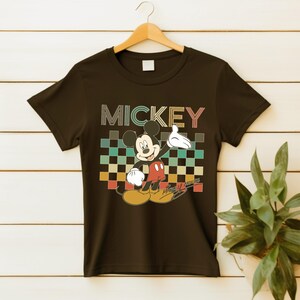 Puede incluir: Camiseta negra con un gr&aacute;fico de Mickey Mouse y la palabra "MICKEY" encima. El dise&ntilde;o presenta a Mickey Mouse en una pose cl&aacute;sica, sobre un fondo a cuadros en tonos turquesa, naranja y verde.