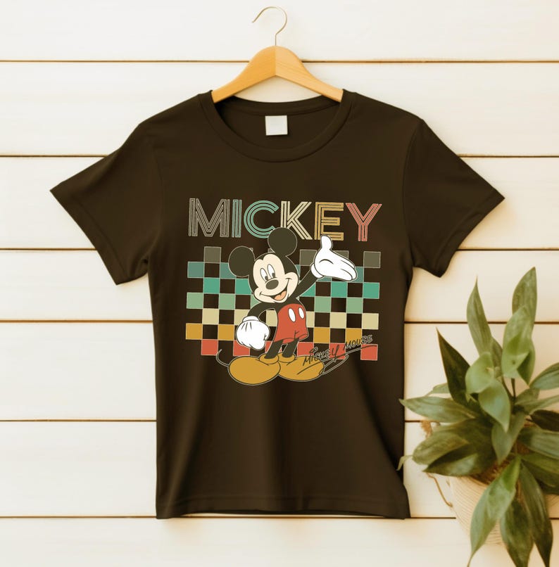 Puede incluir: Camiseta negra con un gr&aacute;fico de Mickey Mouse y la palabra "MICKEY" encima. El dise&ntilde;o presenta a Mickey Mouse en una pose cl&aacute;sica, sobre un fondo a cuadros en tonos turquesa, naranja y verde.