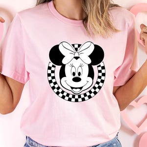 May include: Pink t-shirt with a black and white checkered circle design featuring a cartoon image of Minnie Mouse with a black bow.