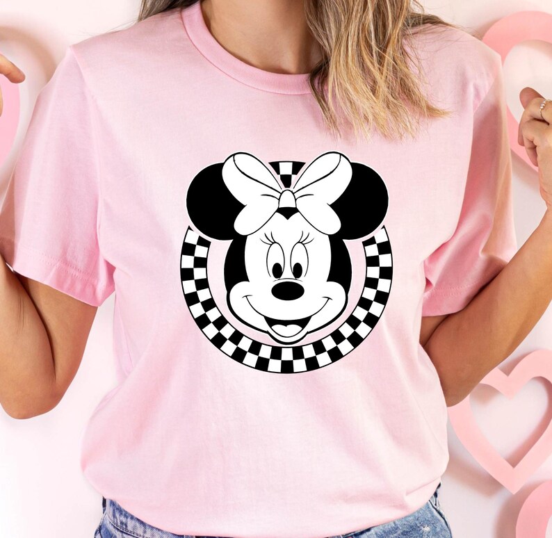 May include: Pink t-shirt with a black and white checkered circle design featuring a cartoon image of Minnie Mouse with a black bow.