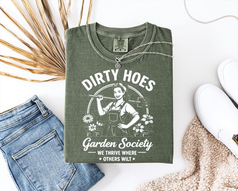 May include: An olive green t-shirt with the text "DIRTY HOES Garden Society" and a graphic of a woman with a hoe. The shirt is folded with a silver necklace, blue jeans, and white sneakers. A dried palm leaf is also in the image.