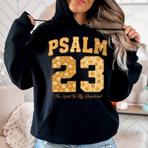 May include: Black hoodie with the text "PSALM 23" in a gold plaid font. The number "23" is in a floral pattern. Below the number is the text "The Lord Is My Shepherd".
