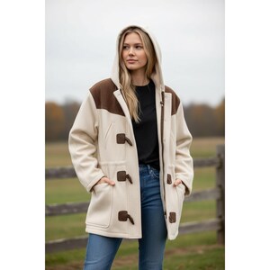 May include: Cream-colored hooded coat with brown shoulder and upper chest accents. The coat features toggle closures and two front pockets. The model is wearing a black shirt and blue jeans. The coat is made of a thick, warm material.