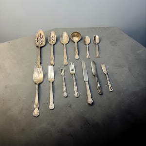May include: An assortment of antique silverware, including spoons, forks, and knives, arranged on a gray surface. The silverware features ornate designs and varying sizes, reflecting a classic aesthetic. The collection includes serving spoons, dinner forks, and butter knives.