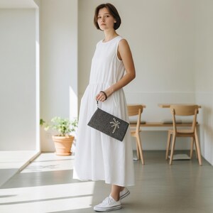 May include: A woman in a white sleeveless dress holds a dark gray clutch with a decorative design. The clutch has a wrist strap and a textured surface. She is wearing white sneakers.