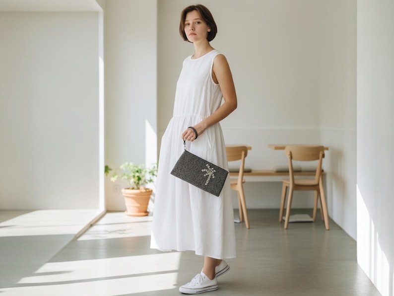 May include: A woman in a white sleeveless dress holds a dark gray clutch with a decorative design. The clutch has a wrist strap and a textured surface. She is wearing white sneakers.