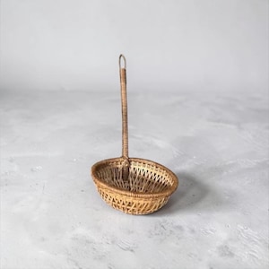 May include: A woven wicker basket with a long handle. The basket is oval-shaped with a handle that curves into a loop at the top. The basket and handle are made of light brown wicker.
