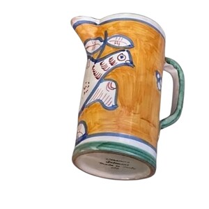 May include: Hand-painted ceramic pitcher with a bird and leaf design. The pitcher features a yellow background with blue, white, and green accents. The handle is green, and the base has text that reads "Ceramiche Salomone made in Italy".