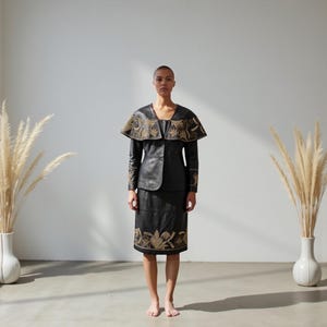 May include: A black leather skirt suit with gold embroidery. The suit includes a jacket with a cape-like collar and a knee-length skirt. The gold embroidery is featured on the collar, sleeves, and skirt hem. The outfit is worn by a person standing in front of a white wall.