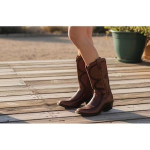 May include: A pair of brown leather cowboy boots with decorative stitching. The boots have a pointed toe and a stacked heel. The boots are worn by a person standing on a wooden deck.