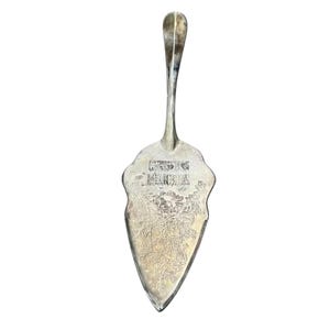 May include: Silver-toned cake server with a decorative handle and a wide, ornate blade. The blade features an engraved design and text. The server is designed for serving cakes and pastries.