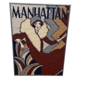 May include: Framed tapestry featuring a woman in a flapper-style dress with the word "MANHATTAN" above her. The artwork uses shades of brown, beige, gray, and blue. The woman is holding a cigarette holder.