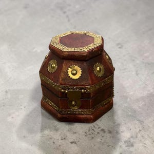 May include: A small, octagonal, vintage-style jewelry box. The box is made of dark red leather with gold-colored metal accents. The lid and sides are decorated with ornate metal details and sunburst-shaped embellishments. The box is closed with a small clasp.