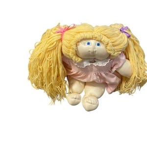 May include: A vintage doll with a soft, plush body and long, yellow yarn hair styled in pigtails. The doll has blue eyes and is wearing a pink dress with a white lace collar. The doll's arms and legs are soft and rounded.