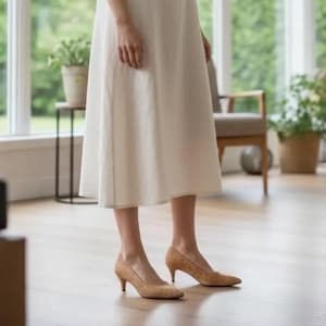 May include: A person wearing a cream-colored dress and tan cork heels with a pointed toe. The heels have a low heel. The background includes a window, plants, and a wooden floor.