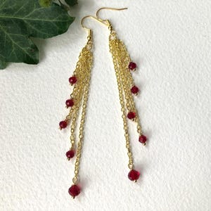 May include: Gold-coloured chain earrings with dangling strands. Each strand features small, round, deep red beads. The earrings have a hook closure. The jewellery is set against a white background.