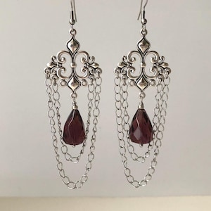 May include: A pair of silver chandelier earrings. Each earring has an ornate filigree top, cascading chain drapes, and a faceted, teardrop-shaped, deep purple gemstone. The earrings are designed for pierced ears.