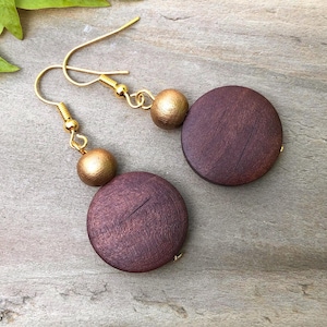 May include: A pair of dangle earrings with large, round, dark brown wooden pendants. Each earring has a smaller, gold-coloured bead and gold-tone ear wires. The earrings are displayed on a light brown surface.