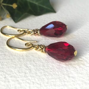 May include: Gold-coloured drop earrings featuring faceted, teardrop-shaped, deep red crystals. The earrings have a decorative gold bead detail near the crystal and a simple gold hook closure. The background is a soft, white surface.