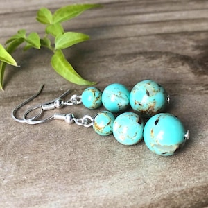 May include: A pair of drop earrings featuring three graduated turquoise-coloured beads with brown matrix veining, suspended from silver-toned metal fishhook wires. The beads increase in size from the top to the bottom of each earring.