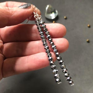 May include: A pair of long dangle earrings featuring two strands of faceted, metallic dark gray beads. The beads are strung on silver-toned wire and attached to silver-toned ear hooks, creating a linear, shimmering design that hangs vertically.