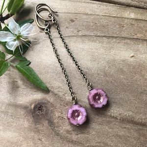 May include: Dangle earrings with pink flower charms. The earrings have a long chain design with antique brass-coloured hooks and chains. The flower charms have a detailed, textured appearance, with a floral theme.