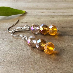 May include: A pair of drop earrings featuring silver-toned metal hooks and three faceted crystal beads each. The beads transition in colour from a pale iridescent champagne to a warm amber and a vibrant golden yellow, creating a gradient effect.