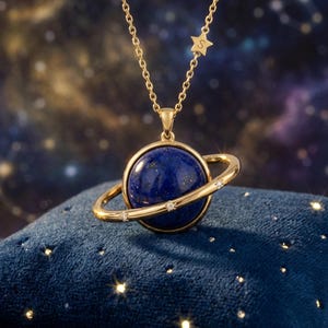 May include: A gold necklace with a planet pendant. The pendant has a blue gemstone sphere and a gold ring with small diamonds. A small star charm with the letter "S" is also included. The necklace is displayed on a blue velvet surface.