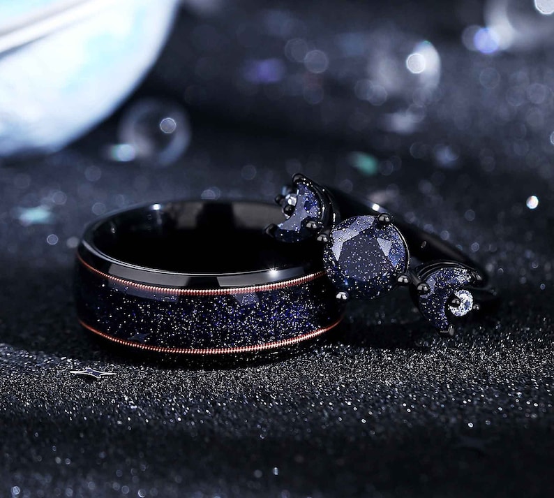 May include: A black wedding band with a copper-coloured band and a black ring with a crescent moon design and a blue sparkly stone.