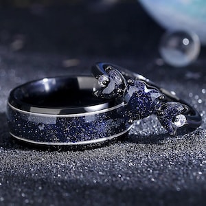 May include: A pair of black wedding bands with a blue starry sky design. The band on the left is a simple band with a blue starry sky design. The band on the right is a heart-shaped band with a blue starry sky design and a small clear stone.