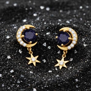Puede incluir: Pendientes dorados con diseño de media luna, una piedra azul oscuro y pequeñas piedras blancas. Cada pendiente tiene una estrella colgante con una sola piedra blanca en el centro. Sobre un fondo oscuro y texturizado.