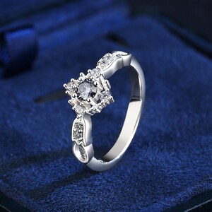 May include: A silver ring with a square-shaped central design and smaller accent stones. The ring has a polished finish, set against a dark blue background. The central design features a central stone surrounded by smaller, sparkling stones.