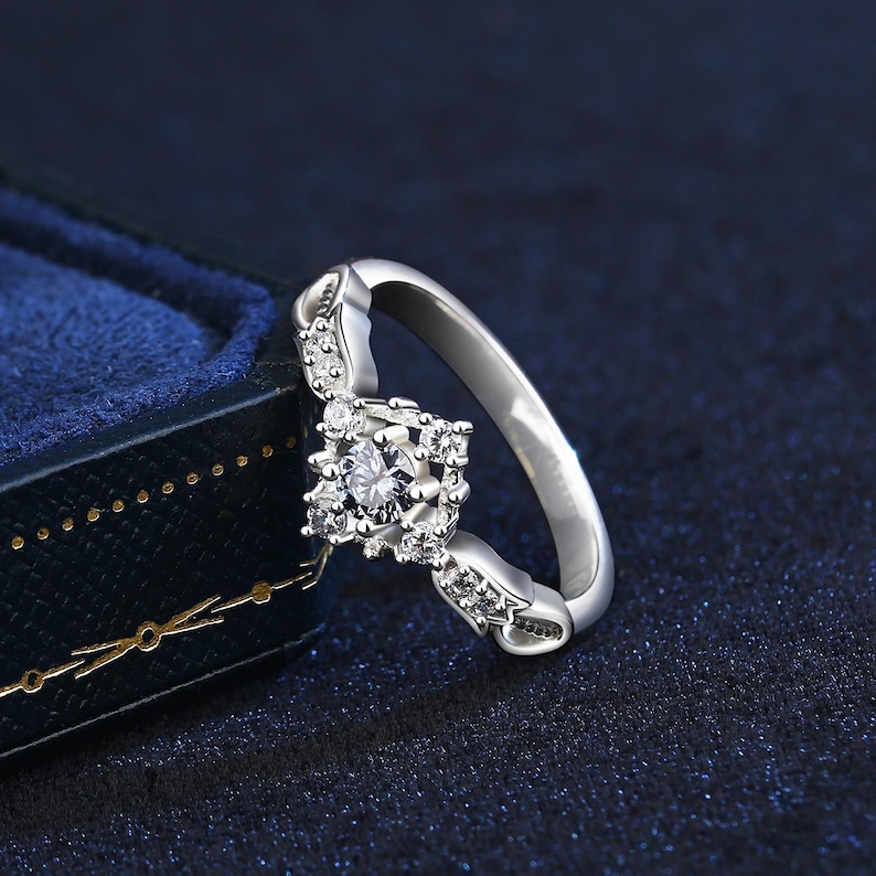 May include: A silver ring with a square, diamond-studded centrepiece. The ring features a central, round diamond surrounded by smaller stones. The band is also adorned with small diamonds. The ring is set against a dark blue background.