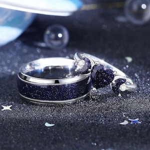 May include: A set of two silver rings with a blue starry sky design. The larger ring features a heart-shaped stone with a crescent moon on each side. The smaller ring has a band with a starry sky design.