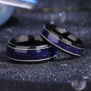 Könnte beinhalten: Zwei schwarze Eheringe mit einem blauen Sternenhimmel-Design. Die Ringe haben einen dünnen silbernen Rand.