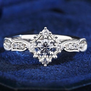 May include: A silver engagement ring with a central round diamond and a square halo design. The ring features smaller diamonds on the band and around the central stone, set against a dark blue background.