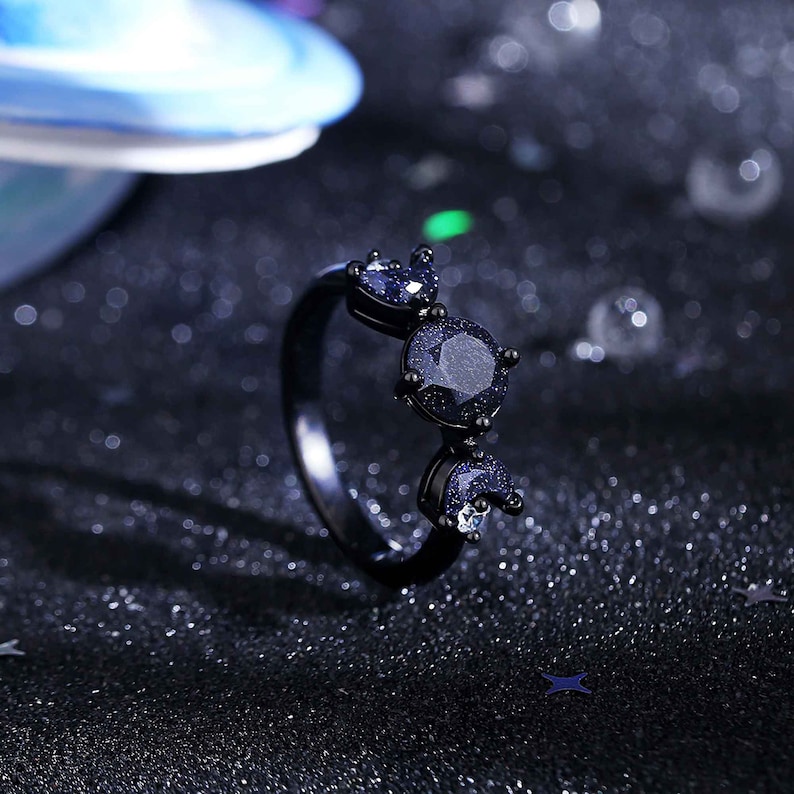 May include: A black ring featuring three blue gemstone accents. The gemstones are round and have a sparkly finish.