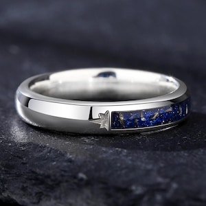 Puede incluir: Un anillo plateado con un acabado pulido. El anillo presenta una incrustación azul oscuro con motas y un diseño en forma de estrella. El anillo está sobre un fondo oscuro.