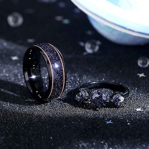 May include: Two black rings with a starry blue design. One ring has a rose gold band. The rings are on a black surface.