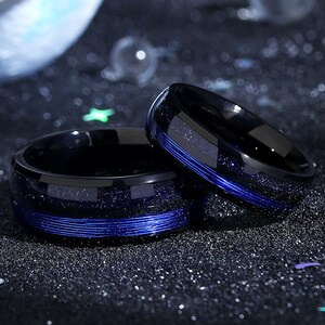 Puede incluir: Dos anillos de carburo de tungsteno negro con líneas azules y un acabado brillante. Los anillos están apilados uno encima del otro.