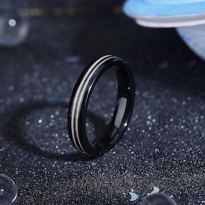 May include: A black ring with a shiny finish, featuring two parallel silver-colored bands. The ring is set against a dark, speckled background, with a blurred object in the background.