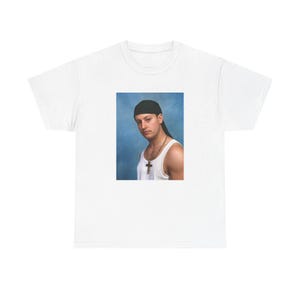 Peut inclure: T-shirt blanc avec un portrait imprimé. Le portrait représente une personne portant un durag noir, un débardeur blanc et un pendentif en forme de croix dorée. Le fond est bleu.