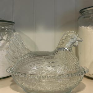 May include: A clear glass hen-shaped dish with a textured surface. The hen sits atop a matching bowl, with a detailed feather design. The dish is likely used for storing eggs or as a decorative kitchen item.