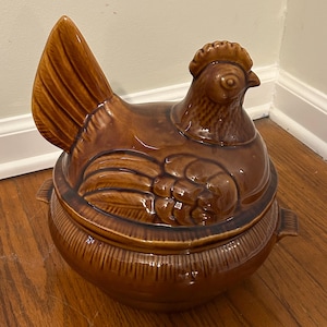 May include: A brown ceramic chicken-shaped casserole dish with a lid. The dish features a detailed design of a chicken with a comb, wattle, and textured feathers. The base has a ribbed pattern and handles on either side.