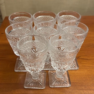 May include: A set of nine clear, patterned glass goblets with square bases. The goblets have a diamond-like pattern and are arranged on a wooden surface. The stemware is suitable for serving beverages.
