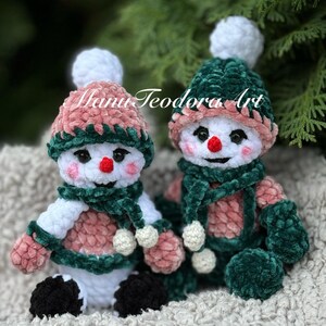 May include: Two crocheted snowman ornaments with white yarn bodies, green velvet hats and scarves, and pink velvet accents. The snowmen are sitting on a white fluffy surface.